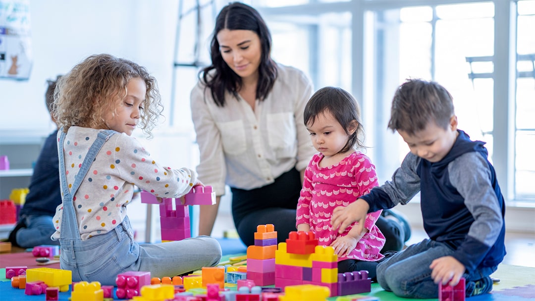 A childcare practitioner wearing a white shirt looks upon a group of children as they play with an assortment of building blocks.
